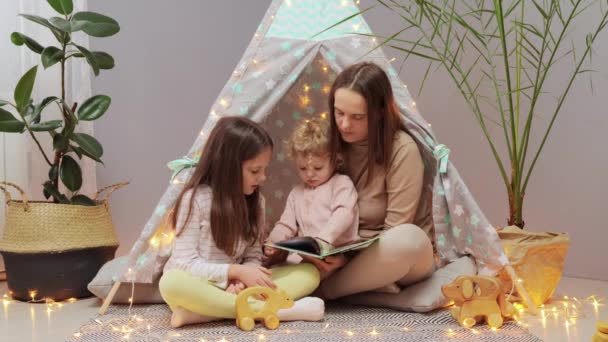 喜ばしい母親と彼女の娘が家内でウィグワムで遊んでいます 読書本 子供を育てる明るい写真を見る面白い妖精のお話 — ストック動画