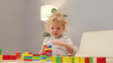 Renkli çocukluk aktiviteleri. Evde şekil sıralaması. Çocuklar için eğitici bir oyun. Bebek bebek, evin içindeki masada tahta geometrik oyuncaklarla oynuyor..