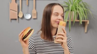 Kafkasyalı kahverengi saçlı kadın çizgili gömlek giyiyor mutfakta oturmuş çizburger yiyor ve sosisli sandviç yiyor abur cuburun tadını çıkarıyor aromat kokuyor..