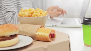Sağlıksız yiyecek, sosisli sandviç, soğan ve bilinmeyen bir kadının laptopta çalıştığı ve işteyken en sevdiği yemekleri yediği yakın plan..