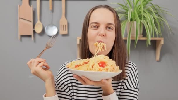 キッチンに座っているストライプのシャツを着た満足した大人の女性 おいしいスパゲッティを食べるおいしいスパゲッティが空腹感を表現 — ストック動画