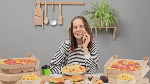トマトとパスタを食べながら 多種多様なジャンクフードを話す電話でテーブルに座っている茶色の髪を持つ笑顔で陽気な陽性の女性 — ストック動画