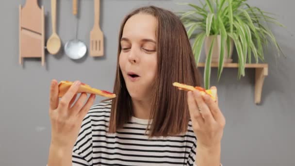 布鲁内特女孩坐在厨房里 满意地吃着比萨饼 看着镜头高兴地吃着披萨 吃着披萨店的快餐 — 图库视频影像