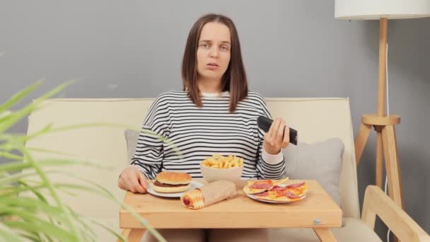 厌倦了的女人坐在沙发上 一边吃着美味的快餐 一边在客厅里看电视 一边吃着外卖小吃 — 图库视频影像