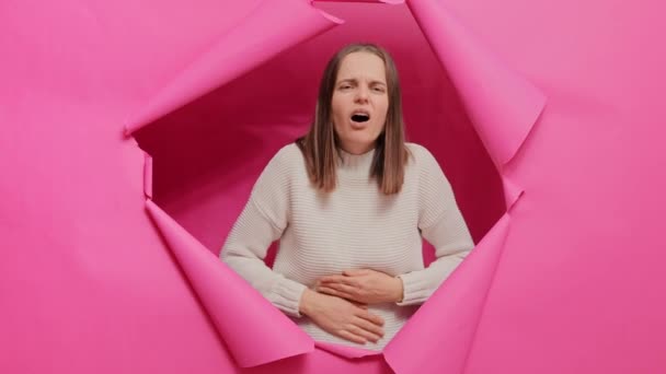 不健康な病気の茶色の毛髪の女性は ピンクの紙の壁の穴を通してポーズセーターを着用し 痛みを感じる彼女の痛みの腹 — ストック動画