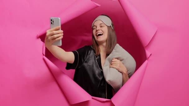 快乐的积极女性 戴着睡眠面具 头戴睡床 手拿着枕头在粉红墙上的纸孔里摆着睡衣 醒来后为社交网站创建了晨间内容 — 图库视频影像