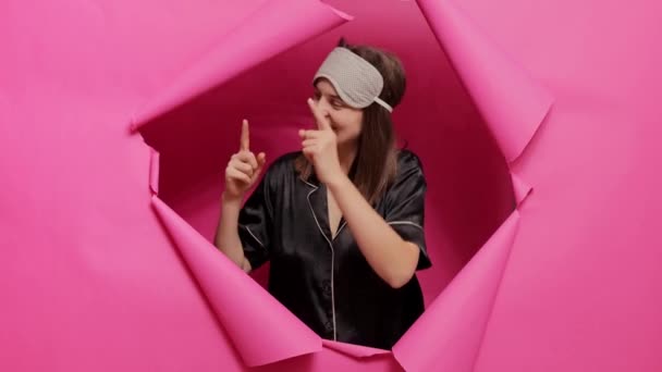 笑逐颜开 笑逐颜开的女士穿过粉红的纸片 身穿睡衣 戴着睡眠面罩 指着旁边的空间广告区 笑逐颜开 — 图库视频影像