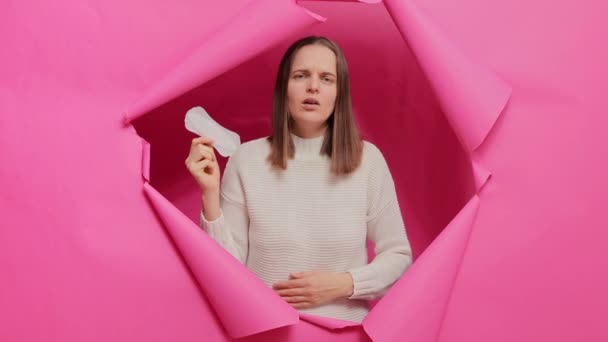 グロミー白人女性は個人的な衛生を気にし 手で衛生パッドを保持し 月経中に痛みを抱え ピンクの紙の壁を突破する悲しい表情をしています — ストック動画