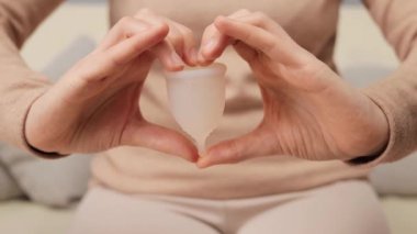 Adet döngüsü rahatlığı. Yeniden kullanılabilir dönem ürünleri. Sıfır adet öncesi bakım. Aybaşı bardağını kalbinin içinde tutan tanınmayan bir kadın ev içinde el hareketi yapıyor.