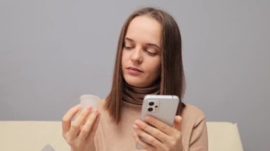 Cep telefonu kullanan ve elinde tekrar kullanılabilir bardak tutan konsantre olmuş genç bir kadın internette alternatif koruma ürünü arama videosunun nasıl kullanılacağını öğreniyor..
