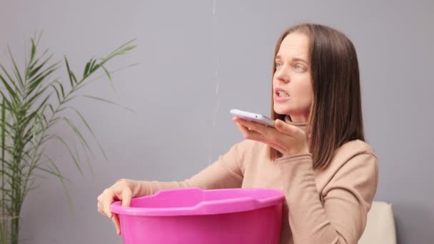 白人不高兴的棕色头发女人拿着脸盆 因为她的屋顶漏水 收集水 打电话给水管工解释她的问题 — 图库视频影像