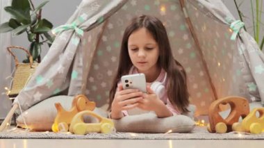 Şaşırmış kahverengi saçlı liseli kız evdeki çadırda uzanıyor akıllı telefon kullanarak internetten ilginç karikatürler arıyor şaşırmış yüzüyle..