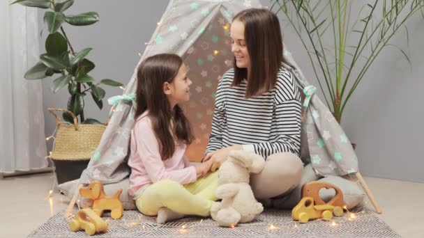 母と娘は一緒にウィグワムに座って 家でリビングルームで抱擁し 笑顔でかわいい小さな女の子をサポートする子供の問題について話します — ストック動画