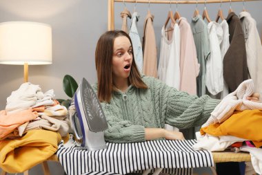 Tekstil bakımı ve hijyen. Şok olmuş, tembel, yetişkin bir kadın. Günlük tişört giyiyor. Ağzı açık bakıyor. Çamaşır yıkadıktan sonra ev işlerini yapmak için ütü yaparken büyük gözleri var.