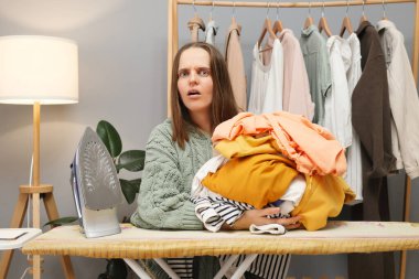 Kafkasyalı kadın kahverengi saçlı, kıyafetlerini ütüleyen evde oturmuş kameraya bakarken fırfırlı çarşaf yığınını kucaklıyor.