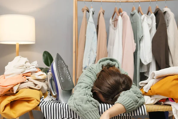 Exhausted adult woman wearing casual shirt falling asleep after long hours ironing clothing being tired of household chores after laundry lying on ironing board