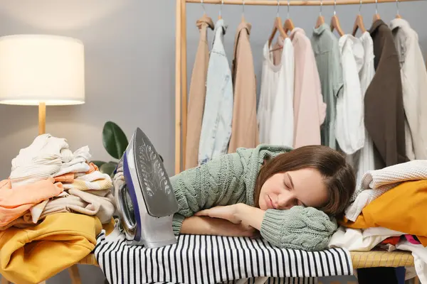 Sleeping lazy brown haired woman wearing knitted shirt ironing clothing while sitting in her wardrobe sleeping on ironing board working with clean apparel at home