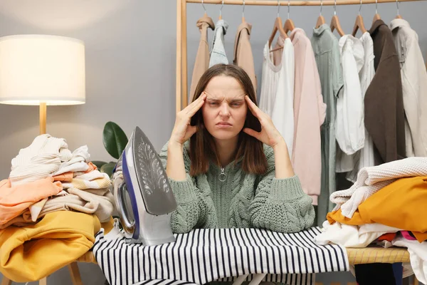 Unhealthy brown haired woman wearing knitted shirt ironing clothing while sitting in her wardrobe having lots household work suffering headache being tired and ill