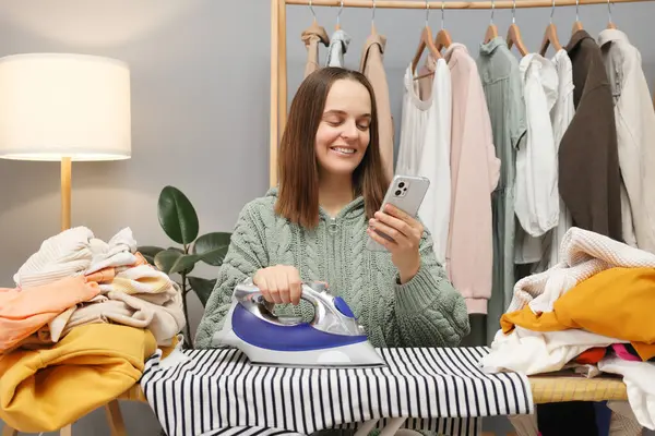 Cheerful brown haired woman wearing knitted shirt ironing clothing while sitting in her wardrobe using mobile phone scrolling online while doing household chores