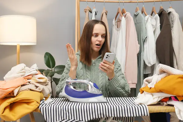 Shocked brown haired woman wearing knitted shirt ironing clothing while sitting in her wardrobe and holding smartphone being surprised posing near ironing board