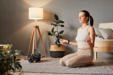 Formda ve çekici bir yaşam tarzı var. Sportif ve ince rutin. Faaliyet için yer. Asana spor salonunda. Yerde oturup yoga yapan güzel bir kadın. İpucu ve tayt, oturma odasında egzersiz yapıyor.