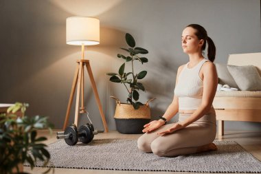 Evde aktif bir yaşam tarzı. Rahatlama ve poz. Sakin ve atletik bir yaşam. İnce bir kadın yerde oturmuş yoga yapıyor. İpucu ve tayt giyiyor. Oturma odasında meditasyon yapıyor.