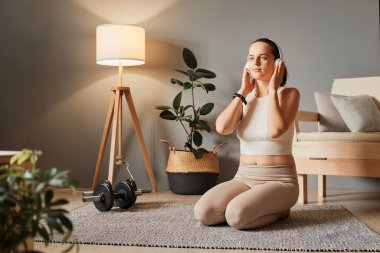 Atletik genç kadın evde egzersiz yaptıktan sonra yerde oturuyor ve kulaklıkla müzik dinliyor. Egzersiz sırasında müzik molası veriyor.