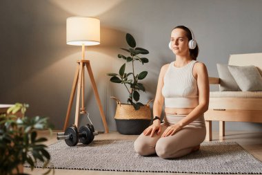 Evde atletik iyileşme. Spor zemin ahengi. Spor kıyafetleri giyen güzel bir kadın antrenmandan sonra dinleniyor oturma odasında oturmuş kulaklık takıyor meditasyonu dinliyor.