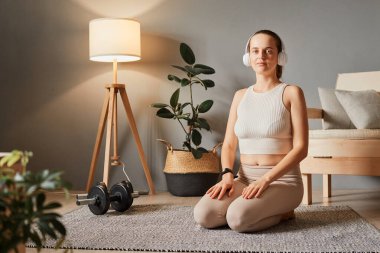 Kapalı alanda spor egzersizi. Kendine güveni tam bir kadın bej spor giyer antrenmandan sonra dinlenir kulaklıkla müzik dinler oturma odasında yerde oturur.