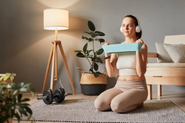Bej spor giyimli, kulaklık takan, kulaklık takan, kollarında elastik bantla egzersiz yapan, kol kasları üzerinde güç antrenmanı yapan, kulaklıkla müzik dinlemenin keyfini çıkaran sportif bir kadın.