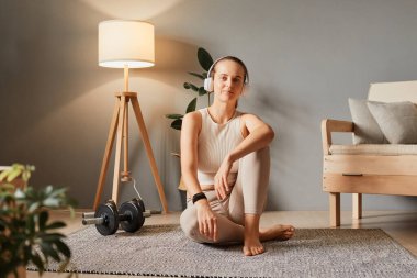 Vücut için sağlık rutini. Zayıf ve formda bir yaşam tarzı. Bakım için kapalı jimnastik. Evde eğitim ve boş zaman. Bej rengi spor giysiler giyen bir kadın antrenman sırasında mola veriyor.
