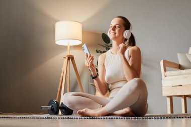 Bej spor giyimli tatmin olmuş bir kadın evde antrenman yaptıktan sonra evde oturup müzik dinliyor ve ev ödevi için en sevdiği çalma listesini dinliyor.