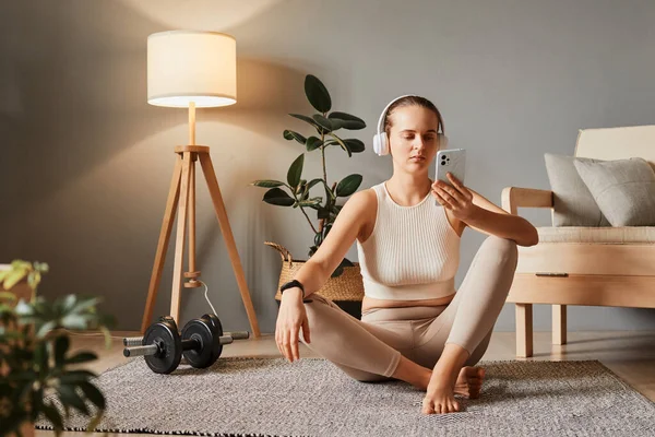 Home gym with technology. Apartment exercise routine. Healthy lifestyle with gadgets. Recreation and fitness at home. Young woman wearing beige tracksuit sitting on floor using mobile cell phone