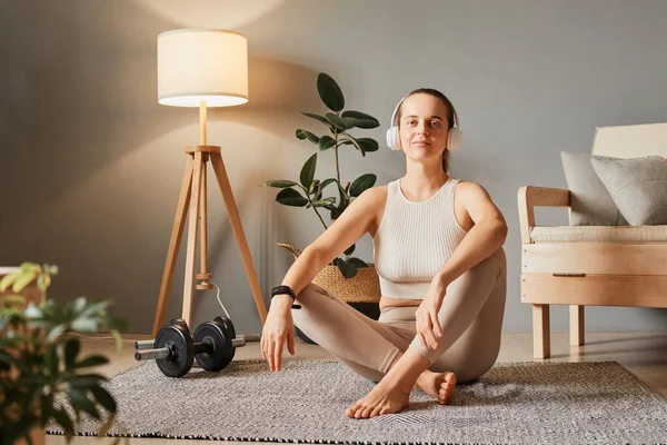Active care for body. Leisure in a sporty lifestyle. Sport and fitness harmony. Positive woman wearing beige sportswear relaxing after home work out sitting on floor in living room looking at camera