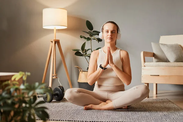 Lifestyle of wellness. Home meditation for health. Happy and healthy living. Fit and sporty routine. Calm woman sitting on the floor practicing yoga wear tip and leggings meditating in living room
