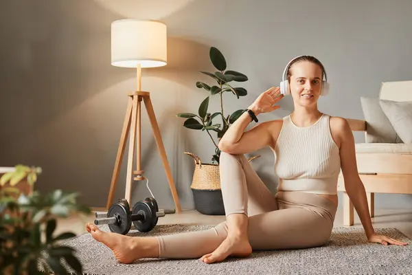 Smiling attractive sporty woman sitting on the floor practicing yoga wear tip and leggings doing exercise in living room listening to music enjoying active home time