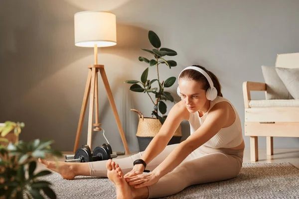 Gymnastics in the house. Fit lifestyle at home. Exercising for health indoors. Pilates for relaxation. Young woman in headphones in sportswear doing fitness stretching exercises at home