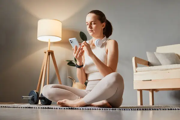Fitness gadget for health. Technology in workout. Online fitness and health. Caucasian adult woman wearing beige tracksuit sitting on floor using mobile cell phone