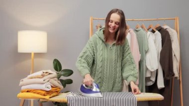 Tekstil için program ütüleniyor. Neşeli, çekici, pozitif, kahverengi saçlı, örgü gömlekli, evinde dans ederken kıyafetlerini ütüleyen bir kadın.