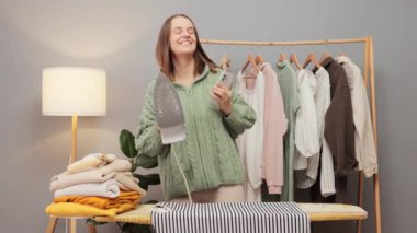 Dans eden, neşeli, kahverengi saçlı, örgü gömlekli genç bir kadın. Evdeki dolabında giysilerini ütülüyor. Ev temizliği keyfi yerindeyken eğleniyor.