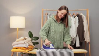 Çalışkan, kahverengi saçlı, örgü gömlekli, elbiselerini ütüleyen, ev işlerini yaparken gülümseyen genç bir kadın.