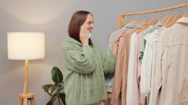 Galeride kıyafet seçen gülümseyen bir kadın cep telefonuyla konuşuyor. Moda mağazasında giysi raflarındaki yeni koleksiyonlara bakıyor.