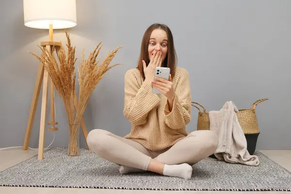 Online betting addiction. Blogger on apps. Amazed Caucasian dark haired woman wearing casual clothing using phone sitting on floor in home interior reading breaking news
