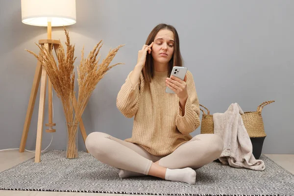 Gadget addiction issue. Pensive Caucasian dark haired woman wearing casual clothing using phone sitting on floor in home interior looking at display with serious face