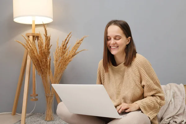 Remote workspace. Online freelancing. Laptop for remote work. Laughing Caucasian woman wearing beige jumper using computer for watching funny videos while sitting on floor in light room