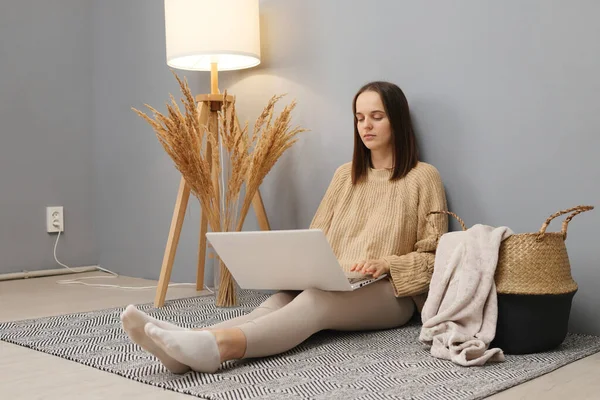 Freelancer\'s workspace. Remote job opportunities. Concentrated calm brown haired woman wearing beige sweater working on computer while sitting on floor in home interior looking at display