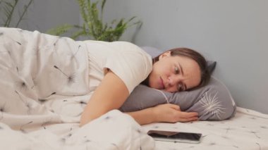 Üzgün, mutsuz, beyaz, akıllı telefonlu bir kadın. Yatak odasında uzanıyor. Akıllı telefondan sıkılıyor ve olumsuz duyguları olan bir şeye bakıyor.