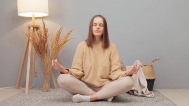 Güzel, genç, beyaz bir kadın evde yoga yapıyor. Ev kıyafetleri içinde. Nilüfer çiçeği pozunda oturmuş meditasyon yaparken sakinleşmeye çalışıyor.