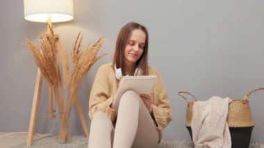 Kahverengi saçlı, güzel bir kadın, bej kazaklı, yerde oturuyor. Organizatörüyle çizim yapıyor. Kadın tasarımcı, evde çizimler çiziyor.