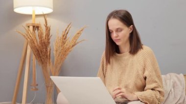 Kapalı mekanda teknik aktiviteler. Uzaktan kumanda. Oturma odasında bilgisayar ekranına bakıp esnerken bej kazak giyen yorgun bir kadın.